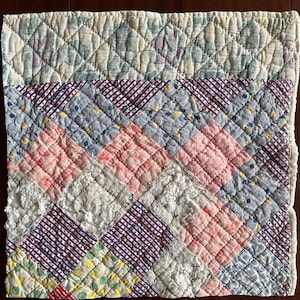 May include: A patchwork quilt with a variety of patterned fabrics in shades of pink, blue, white, and green. The quilt features a diamond pattern and is hand-stitched. The edges are finished with a white trim. The quilt is a vintage item.
