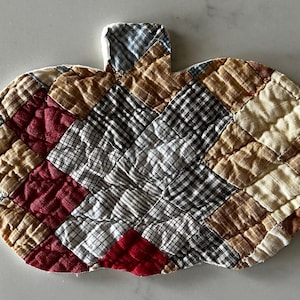 May include: A pumpkin-shaped quilted fabric mat featuring a patchwork design of small squares in various patterns and colors, including red, black and white gingham, tan, and brown plaid. The quilted texture is visible across the entire surface.