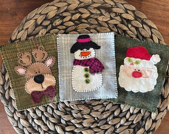 May include: Three Christmas-themed felt squares: a reindeer with a burgundy bow tie, a snowman with a black hat and purple scarf, and Santa Claus with a red hat. Each square is stitched with white thread.