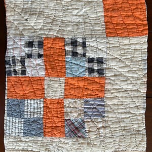 May include: Handmade patchwork quilt featuring a grid pattern of squares in various colors and patterns. The quilt incorporates orange, white, blue, and plaid fabrics. The quilt has a textured, quilted surface and a vintage aesthetic.