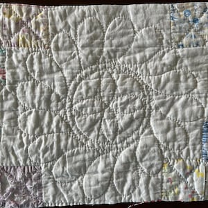 May include: A white, quilted textile with a central floral design. The quilt features a repeating pattern of large, petal-shaped elements. The corners are adorned with small, colorful patchwork squares, adding a touch of contrast.