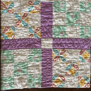 May include: A vintage quilt with a patchwork design. The quilt is made of white fabric with squares of green, purple, and floral print fabric. The quilt is quilted with a simple stitch pattern.