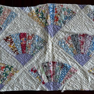 May include: A rectangular quilt featuring a repeating fan pattern. Each fan is composed of multiple fabric strips in various floral and geometric prints, anchored by a solid light purple base. The background is white with quilted grid lines.