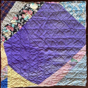 May include: A patchwork quilt with a large central diamond in shades of purple, quilted with horizontal lines. The quilt's edges feature various patterned fabrics, including floral, plaid, and geometric designs. The quilt is square and appears handmade.