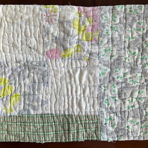 May include: A handmade quilt with a patchwork design. The quilt features various fabric patterns, including floral prints in pink, yellow, and gray, and a green patterned border. The quilt is made of soft, textured fabric.