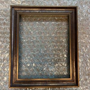 May include: A dark brown wood picture frame with a gold-coloured inner edge. The frame is on a white bubble wrap background.