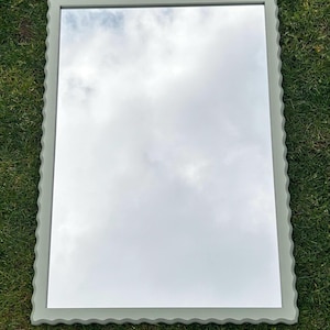 May include: A large, rectangular mirror with a scalloped, light grey frame. The mirror reflects a cloudy sky.