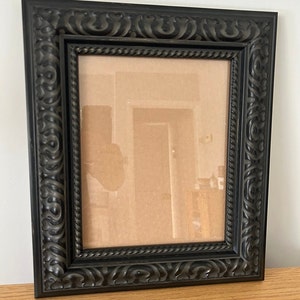May include: A black picture frame with an ornate, carved design. The frame has a rectangular shape and a dark finish. The inner area is empty, ready for a photo or artwork. The frame is sitting on a wooden surface.