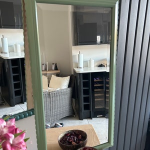 May include: A green framed mirror with a scalloped edge, reflecting a room with a wicker chair and black cabinets. The mirror is sitting on a wooden surface with two bowls of dried rose petals.