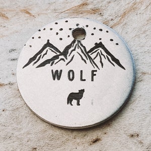 May include: A circular silver-toned metal tag featuring a black engraved design of three mountain peaks under a starry sky. Below the mountains, the word WOLF is printed in capital letters, followed by a small silhouette of a howling wolf.