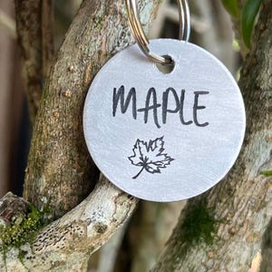 Könnte beinhalten: Ein silbernes Metallschild mit dem Wort "MAPLE" in schwarzen Lettern graviert. Unter dem Wort ist auch ein kleines Ahornblatt graviert.