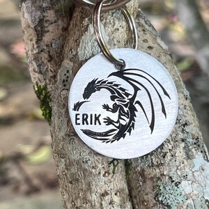 May include: Silver dog tag featuring a black dragon design with the name "ERIK" engraved on it.