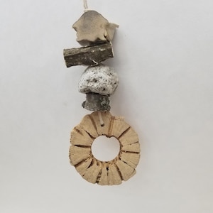 May include: A hanging ornament composed of a vertical stack of natural elements including a light-colored shell, a dark wooden twig, a porous white stone, a small dark wood segment, and a circular, textured tan wood slice at the base.