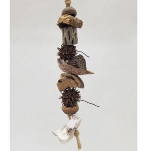 May include: A hanging crabitat toy made of natural materials. The toy features various textures and shapes, including wood, seed pods, and a shell, strung together with twine. The toy is designed for small pets and has the text "Crabiturals" at the bottom.