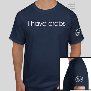 May include: A navy blue t-shirt with the text "i have crabs" printed in white on the front. The shirt has a white spiral design on the sleeve. The shirt is available in a variety of colors, including purple, royal blue, red, sport grey, metro blue, black, navy, and blue dusk.