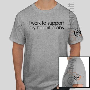 May include: A heather gray short-sleeve t-shirt featuring the black text "I work to support my hermit crabs" printed on the chest. The left sleeve displays a small black and orange spiral shell graphic, which is also visible in the inset image.