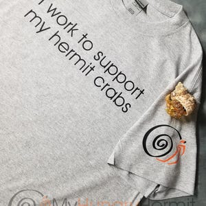 May include: A heather gray t-shirt featuring the black text I work to support my hermit crabs. The sleeve displays a black spiral shell graphic with orange legs, and a small hermit crab in a shell rests on the fabric near the sleeve.