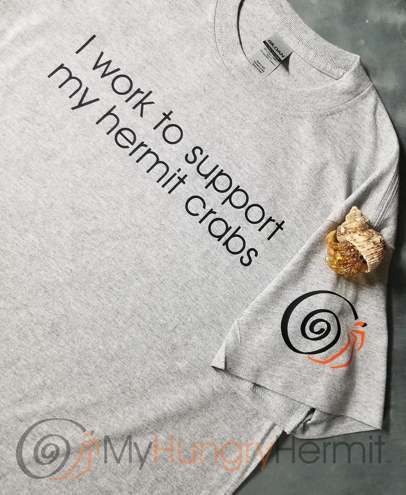 May include: A heather gray t-shirt featuring the black text I work to support my hermit crabs. The sleeve displays a black spiral shell graphic with orange legs, and a small hermit crab in a shell rests on the fabric near the sleeve.