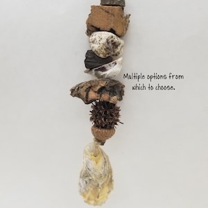 May include: A vertical hanging ornament composed of natural elements including a wooden block, a textured stone, a dark shell, a piece of driftwood, a spiky seed pod, an acorn, and a large oyster shell on a jute rope. Text reads: Multiple options from which to choose.