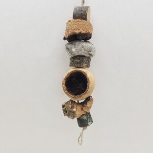 May include: A vertical hanging toy composed of natural elements strung on a light-colored cord. The arrangement includes small wooden segments, a textured grey stone, and a hollow, round, tan-colored seed pod with a dark interior cavity.