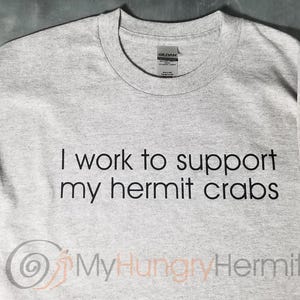 May include: A light gray crew neck t-shirt featuring the black text I work to support my hermit crabs. The shirt includes a graphic of a spiral shell and a small hermit crab on the sleeve, with the text MyHungryHermit printed at the bottom.