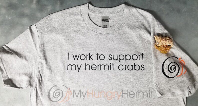 May include: A light gray crew neck t-shirt featuring the black text I work to support my hermit crabs. The shirt includes a graphic of a spiral shell and a small hermit crab on the sleeve, with the text MyHungryHermit printed at the bottom.