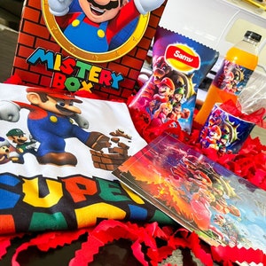 Puede incluir: Una caja de regalo roja y blanca con las palabras "Mystery Box" en ella. La caja presenta una imagen de dibujos animados de Mario de la serie de videojuegos Super Mario. La caja está rodeada de papel de seda rojo y otros artículos con temática de Mario, como una camiseta, una bolsa de patatas fritas, una caja de zumo y una taza.