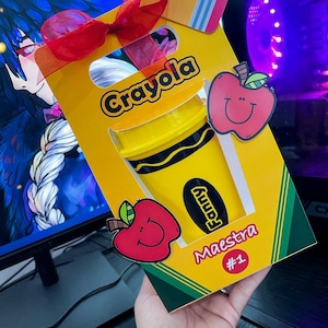 Puede incluir: Una caja de regalo amarilla con temática Crayola con una cinta roja, con un gráfico de crayón amarillo. La caja incluye una pegatina de manzana roja sonriente y el texto "Fanny Maestra #1".