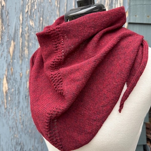 May include: A deep red knitted cowl scarf on a mannequin. The scarf features a textured finish and an openwork design along the edge. The cowl is designed to be worn around the neck and is made of a soft, warm material.