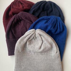 Puede incluir: Cinco gorros de punto de diferentes colores: burdeos, azul oscuro, azul claro, gris y gris claro. Los gorros están apilados uno encima del otro.