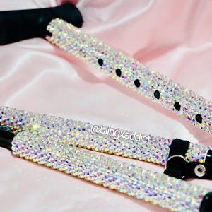 May include: Two bedazzled canes with black handles and straps. The canes are covered in iridescent crystals, reflecting various colors. The background is a soft pink satin fabric. The text "@blingxaddict" is visible.