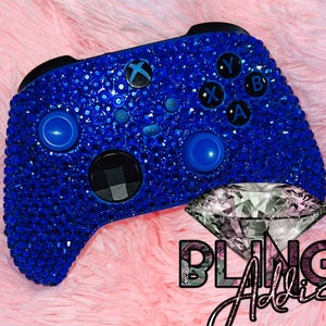 May include: A blue Xbox controller covered in sparkling blue rhinestones. The controller has a black diamond-shaped button in the center and the letters X, Y, A, and B are visible on the top of the controller. The text "BLING Addict" is visible in the lower right corner of the image.