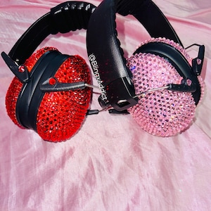 May include: Two sets of bedazzled earmuffs. One set is red, the other is pink. Both have black headbands and ear cups. The earmuffs are covered in small, sparkling rhinestones. The background is a light pink, silky fabric.