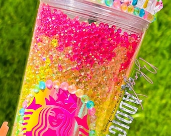 May include: A clear plastic tumbler with a rainbow of colorful crystals inside. The tumbler has a white lid with a rainbow swirl design and a yellow M&M candy on top. The tumbler has a pink Starbucks logo on the front and the text "ADDICT CUSTOM CRYSTAL ACCESSORIES" on the side.