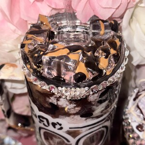 May include: A clear tumbler with a black and white animal print design and a silver glitter rim. The tumbler is filled with ice and a brown liquid, and has the word "COFFEE" written on the side.