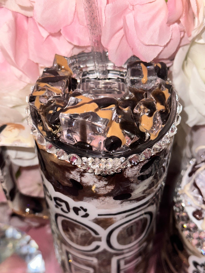 May include: A clear tumbler with a black and white animal print design and a silver glitter rim. The tumbler is filled with ice and a brown liquid, and has the word "COFFEE" written on the side.