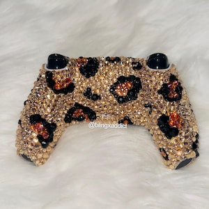 May include: A bedazzled video game controller with a leopard print design. The controller is covered in gold, black, and red rhinestones. The text "@blingxaddict" is visible on the bottom of the controller.
