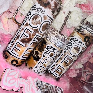 May include: Three brown and white coffee tumblers with a black and white "Coffee" design and rhinestones. The tumblers are on a pink surface with a pink feather boa and a sign that says "Bling" and "Custom Crystal Accessories".