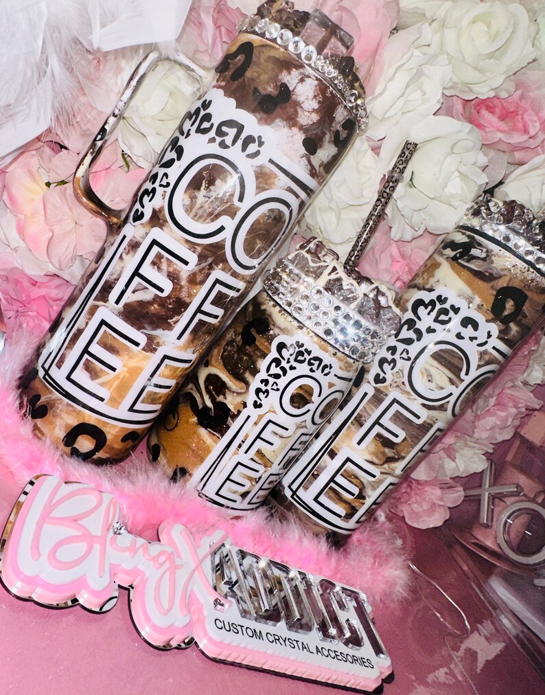 May include: Three brown and white coffee tumblers with a black and white "Coffee" design and rhinestones. The tumblers are on a pink surface with a pink feather boa and a sign that says "Bling" and "Custom Crystal Accessories".