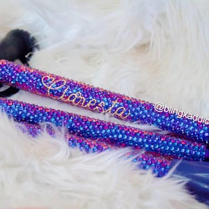 May include: A walking cane featuring a black handle and a shaft covered in a dense pattern of sparkling blue and magenta rhinestones. The name "Claresta" is written in gold script on the shaft, and the text "@blingxaddict" is visible nearby.