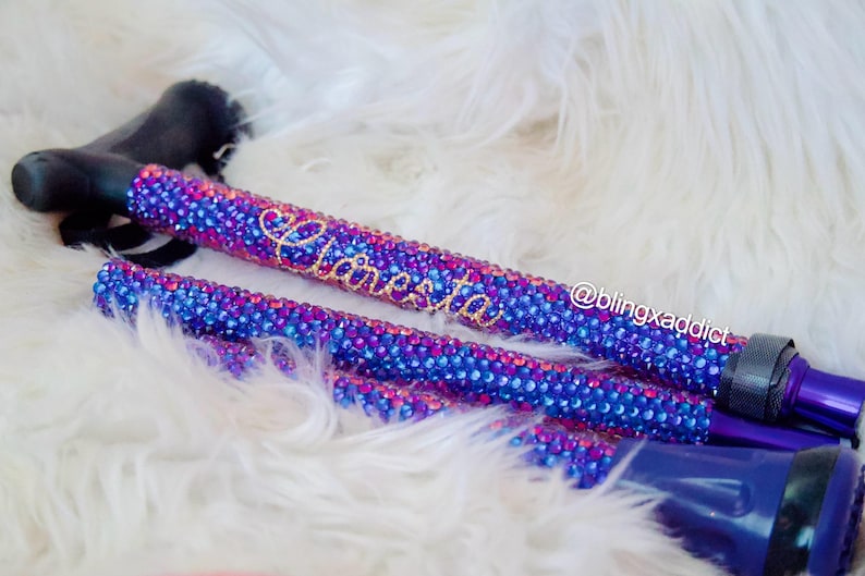 May include: A walking cane featuring a black handle and a shaft covered in a dense pattern of sparkling blue and magenta rhinestones. The name "Claresta" is written in gold script on the shaft, and the text "@blingxaddict" is visible nearby.