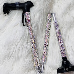 Puede incluir: Un bastón plegable negro con asa y correa para la muñeca. El eje está cubierto de pedrería iridiscente. El bastón está parcialmente plegado, revelando su diseño segmentado. Diseñado para apoyo y movilidad.