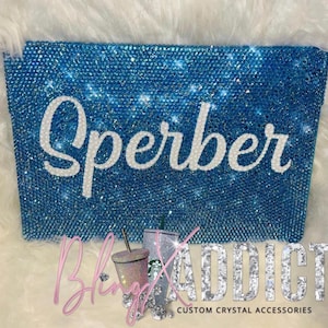 May include: A blue, crystal-covered clutch with the name "Sperber" in white script. The clutch is embellished with sparkling crystals. The image also includes crystal-covered tumblers and the text "Bling Addict Custom Crystal Accessories."