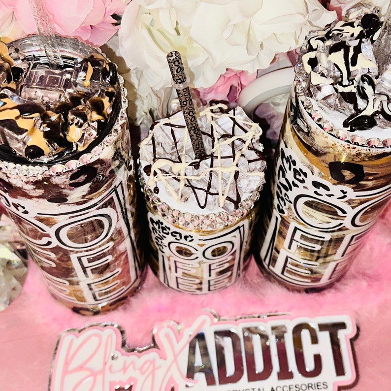 May include: Three coffee tumblers with a brown and white leopard print design. The tumblers have a clear plastic lid and a straw. The tumblers are decorated with white and brown swirls and rhinestones. The tumblers are sitting on a pink surface with a sticker that says "Bling Addict Crystal Accessories".