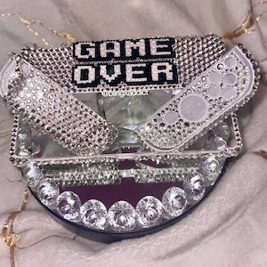 May include: A white Nintendo Switch console and two controllers covered in clear rhinestones with the words "GAME OVER" in black and white pixelated letters. The console is on a mirrored base with a rhinestone border.