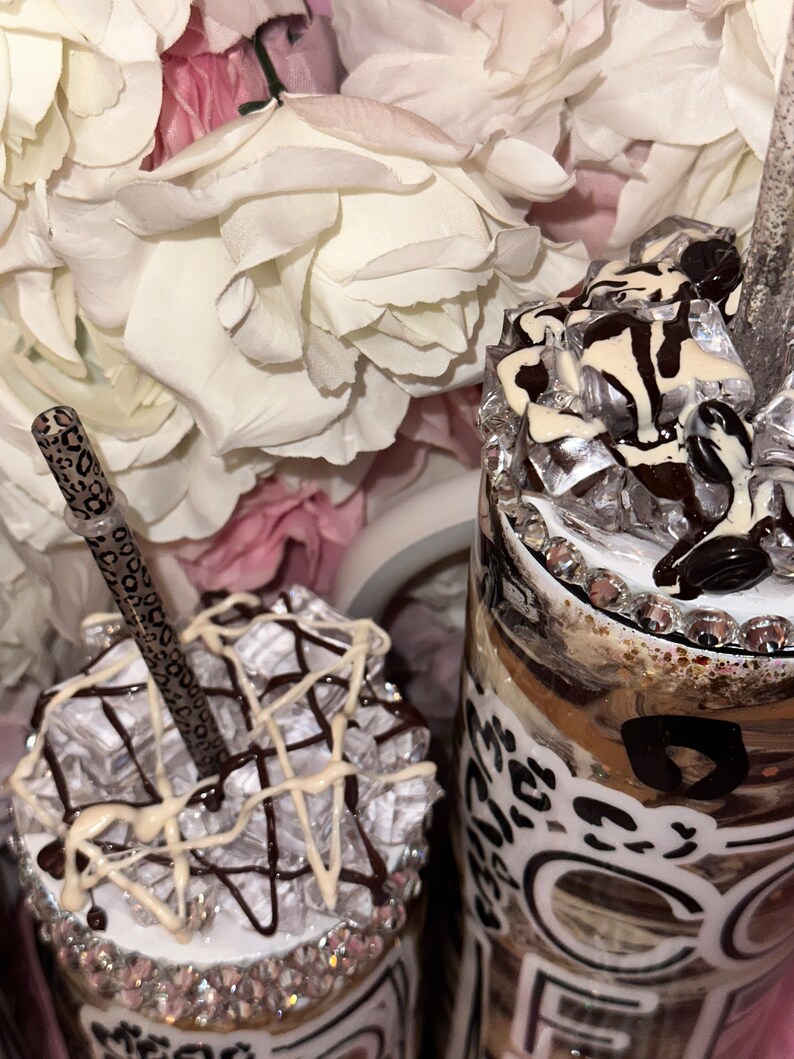 May include: Two personalized coffee tumblers with a brown and white leopard print design. The tumblers are decorated with white and brown chocolate and topped with clear plastic ice cubes. One tumbler has a straw with a leopard print design.