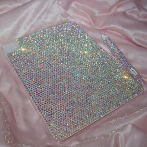 May include: A rectangular tablet case and a matching stylus pen, both fully covered in iridescent, multi-colored rhinestones that create a sparkling effect. The text @blingxaddict is printed in white on the surface of the tablet case.