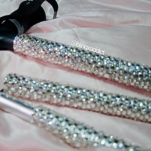 May include: A folding walking cane featuring a black handle with a black wrist strap and a shaft covered in clear, reflective rhinestones. The text @blingxaddict is printed in white near the top of the shaft, which is composed of three segments.