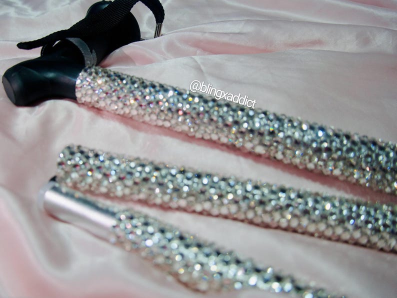 May include: A folding walking cane featuring a black handle with a black wrist strap and a shaft covered in clear, reflective rhinestones. The text @blingxaddict is printed in white near the top of the shaft, which is composed of three segments.