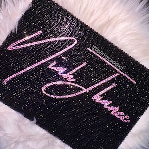 May include: A black clutch bag covered in small, sparkling black crystals. The words "Rich Babe" are spelled out in pink crystals across the front. The Instagram handle @blingxaddict is also visible.